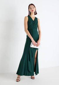 Rochie lungă de culoare verde smarald, cu decolteu adânc în V, design înfășurat, fante laterale și material fluid. Asortată cu o poșetă argintie și sandale negre cu barete.