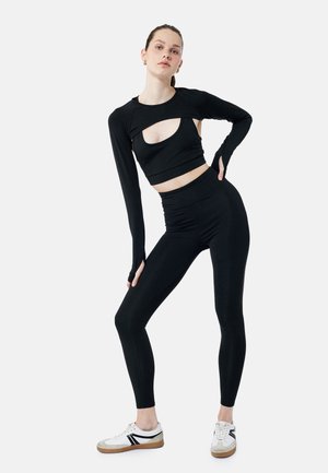 Sort tætsiddende crop top med lange ærmer og udskæringsdesign, parret med højtaljede sorte leggings og afslappede hvide sneakers.
