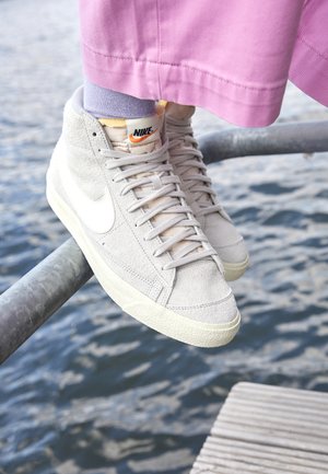Person iført lysegrå Nike høje sneakers og lavendelfarvede sokker, siddende på et metalgelænder ved vandet, med synlige lyserøde bukser.