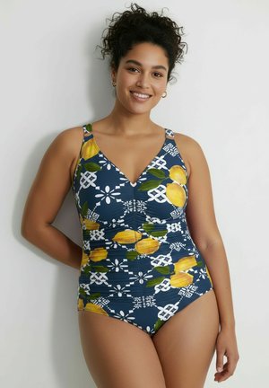Uśmiechnięta kobieta z kręconymi włosami nosi niebieski jednoczęściowy kostium kąpielowy z żółtymi owocami i białymi geometrycznymi wzorami, stojąca przy jednolitej ścianie.