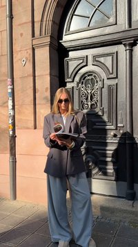 Osoba v přehnaně velkém šedém saku a širokých kalhotách čte knihu, zatímco stojí před tmavými zdobenými dveřmi s obloukovým oknem.