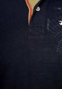 Marineblaues Polohemd mit kontrastierendem gelben Kragen, roten Akzenten und einem weißen Grafikdruck auf der Brust, aus strukturiertem Baumwollmaterial gefertigt.