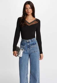 Zwart langemouwen top met een doorschijnende, gestippelde mesh halslijn, gecombineerd met high-waisted blauwe jeans en een metalen zilveren clutch.