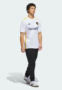 Vit sporttröja med diagonala ränder, gula detaljer och en logotyp. Matchad med svarta cargo-byxor och vita sneakers med svarta ränder.