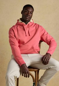 Homme assis portant un sweat à capuche corail sur une chemise beige à col et un pantalon beige clair, devant un fond uni beige.