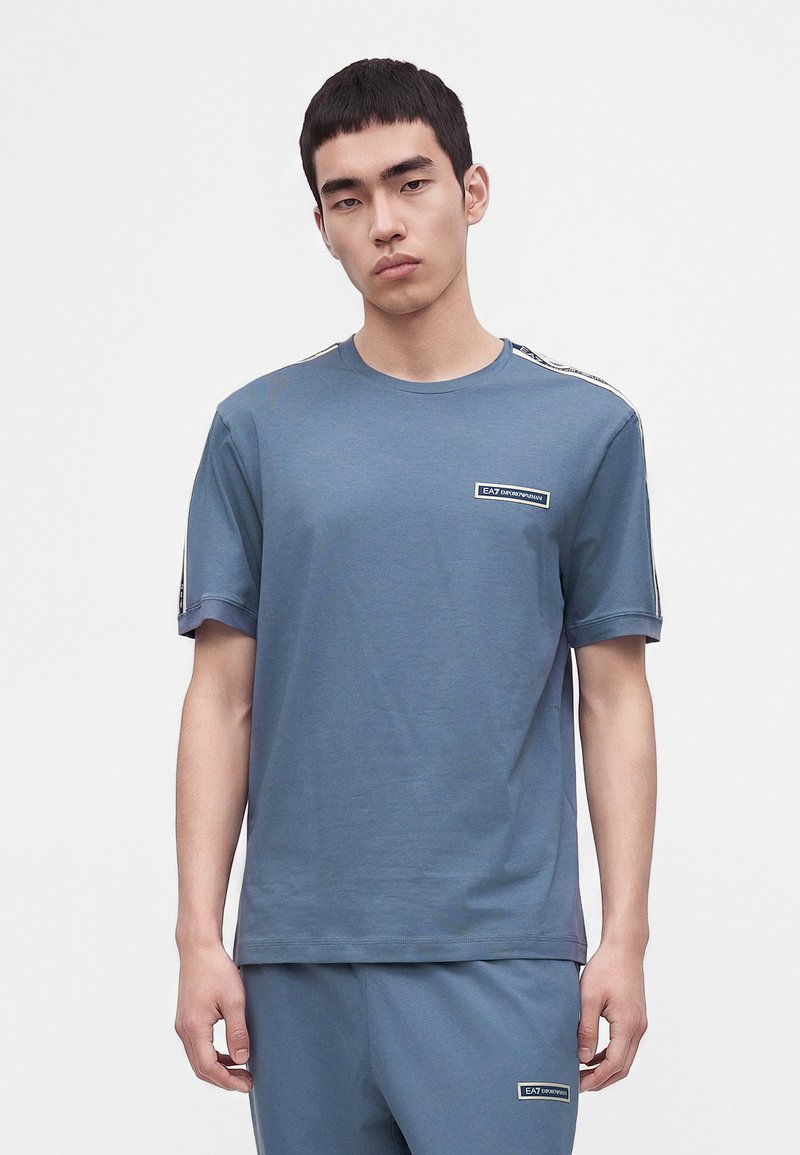 Νέος άνδρας που φοράει μπλε μπλουζάκι EA7 Emporio Armani και ταιριαστό παντελόνι στέκεται μπροστά σε λευκό φόντο.