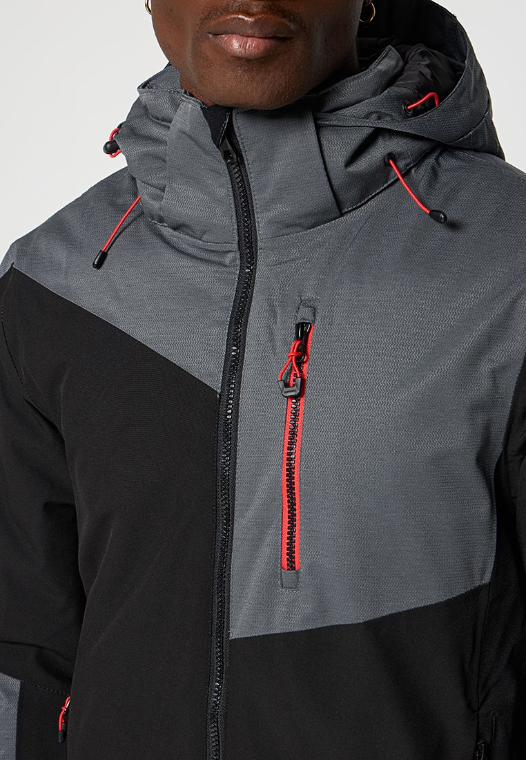Hombre con chaqueta negra y gris de cuello alto, tiradores de cremallera rojos y capucha con cordones rojos.