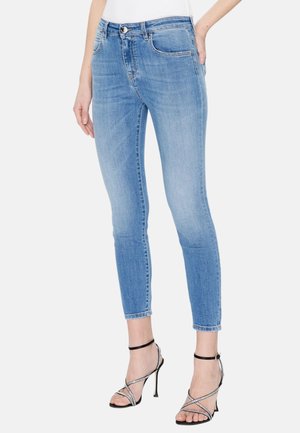 Femme portant un jean skinny bleu clair et des sandales noires à talons hauts avec des brides, debout avec une main dans la poche.