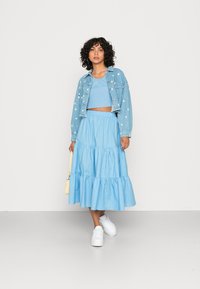 Jaqueta de denim azul claro com tachas decorativas, top cropped canelado a condizer e uma saia longa em camadas. Ténis brancos e uma pequena mala amarela.
