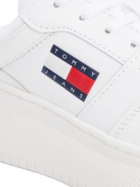 Sapatilha branca com textura de couro suave, apresentando um logótipo retangular proeminente em azul, branco e vermelho na lateral e uma sola de borracha texturizada.