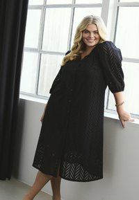 Robe noire midi avec manches bouffantes et fermeture à boutons sur le devant. Fabriquée en tissu texturé avec un motif en chevrons. Comprend une fente sur le côté et un tissage ouvert.