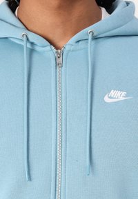 Lyseblå Nike zip-up hettegenser med snøring, med en metallisk glidelås og hvit Nike-logo på venstre bryst. Myk tekstur.