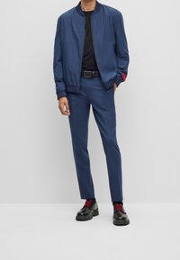 Man in een op maat gemaakte blauwe broek met bijpassende bomberjack, zwart shirt, zwarte riem en zwarte schoenen met rode veters, staande tegen een effen achtergrond.