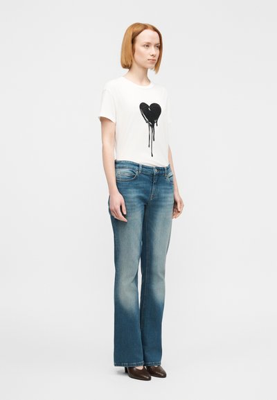 Λευκό μπλουζάκι με μαύρο γραφικό καρδιάς και σταγόνες· συνδυασμένο με ανοιχτόχρωμο τζιν καμπάνα. Το μοντέλο στέκεται όρθιο με κοντά μαλλιά.