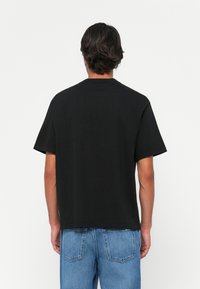 Hombre con cabello oscuro que lleva una camiseta negra de manga corta lisa y jeans azules, de pie frente a un fondo blanco con la espalda a la cámara.