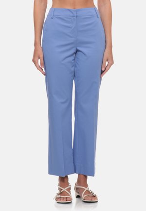 Donna con pantaloni blu a vita alta e taglio corto con tasche laterali e sandali bianchi con tacco e cinturini su sfondo bianco.