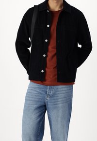 Homme portant une veste en velours côtelé noire boutonnée sur une chemise couleur rouille et un jean bleu clair, portant un sac à bandoulière noir.