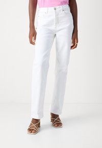 Jeans de mezclilla blancos de tiro alto con piernas rectas y cierre de botón en la parte frontal. Combinados con sandalias de tacón cuadrado beige con tiras.