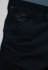 Donker marineblauwe katoenen broek met een gladde textuur. Kenmerken zijn een gebogen zakontwerp en merknaam op de tailleband.