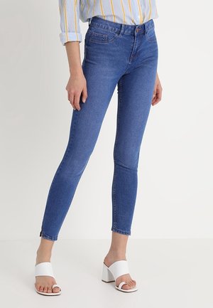 Frau trägt blaue Skinny-Jeans, ein hellblaues Hemd mit gelben vertikalen Streifen und weiße offene Sandalen mit Blockabsatz.