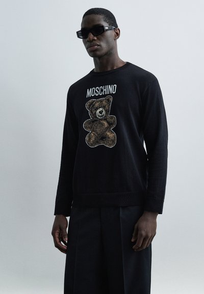 Homme portant un pull noir avec un graphisme d'ours en peluche et le texte « MOSCHINO », un pantalon noir à jambes larges, et des lunettes de soleil noires sur un fond gris.