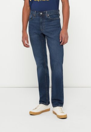 Man draagt donkere blauw recht gesneden spijkerbroek, witte sneakers met bruine zolen en een donkerblauw shirt met gele tekst, staande tegen een witte achtergrond.