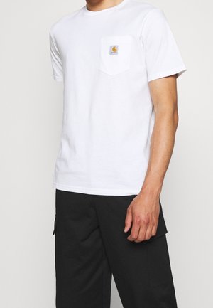 Homme portant un t-shirt blanc uni à manches courtes avec une poche poitrine et un pantalon cargo noir, debout devant un fond uni.