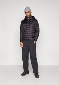 Casaco preto acolchoado com capuz, fecho de correr e bolsos com fecho, combinado com calças cargo pretas e calçado escuro, contra um fundo neutro.