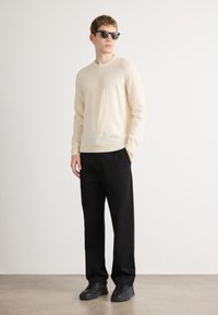 Svetlý krémový sveter s dlhými rukávmi, čierne voľné nohavice a čierne tenisky. Model má na sebe tmavé slnečné okuliare. Minimalistický štýl.