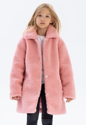 Cappotto in pile rosa con una texture morbida, colletto grande e chiusure a scatto frontali, indossato sopra una maglietta bianca e pantaloni neri.