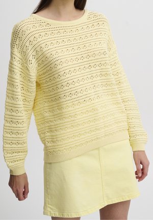 Gul strikket sweater med en struktureret, åben mønster og ribbede manchetter, parret med en matchende gul denimnederdel.