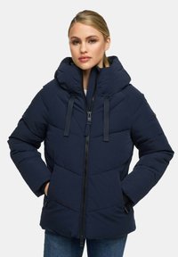 Vrouw in een donkerblauwe gewatteerde jas met een hoge kraag en kap, met de handen in de zakken, tegen een lichte effen achtergrond.