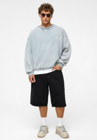 Lichtgrijze oversized sweatshirt met ribgebreide boorden, gecombineerd met zwarte denim shorts en grijze sneakers. Accessoires zijn onder meer zonnebril en gelaagde kettingen.