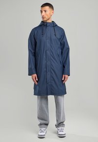 Navy waterproof raincoat with a hood, snap buttons, two side pockets, and adjustable drawstring. Worn over gray pants and sneakers.