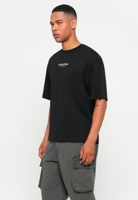 Zwarte t-shirt met korte mouwen en een ronde hals, met witte tekst "STUDIO EDITION" op de voorkant. De stof lijkt soepel en lichtgewicht.