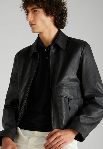 Chaqueta de cuero negra con un cuello con solapas, cremallera frontal y un bolsillo en el pecho. Textura suave y corte entallado. Polo negro debajo.