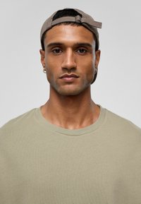 Beige katoenen t-shirt met een ronde hals. Het onderwerp draagt een lichtbruine pet en heeft een kleine oorbel. Gladde textuur, neutrale tinten.