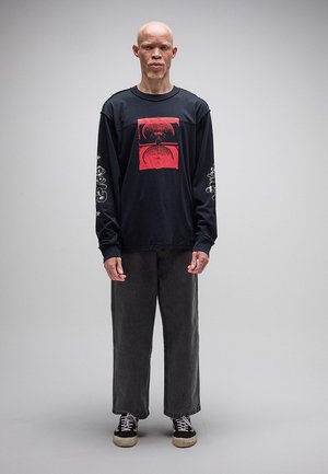 Moški z albinizmom stoji oblečen v črno dolg rokav s rdečim potiskom, ohlapne temne hlače in črne superge na sivi podlagi.