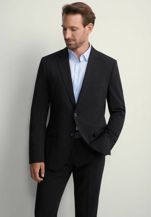 Czarna marynarka w stylu klasycznym z klapami, zapinana na jeden guzik, z dwiema przednimi kieszeniami, noszona na jasnoniebieskiej koszuli. Gładka tekstura.