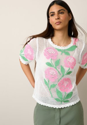 Femme aux longs cheveux foncés portant un haut en maille blanc avec des motifs de fleurs roses et de feuilles vertes et un pantalon vert, debout, les mains derrière le dos.