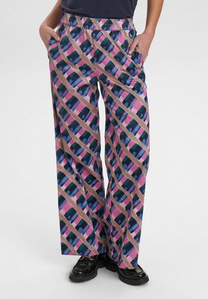 Persona que lleva pantalones anchos y sueltos con rayas geométricas diagonales multicolor en rosa, azul, verde y beige, con las manos en los bolsillos y zapatos negros.