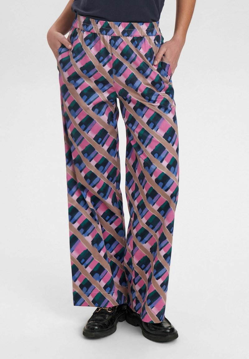 Personne portant un pantalon ample à jambes larges avec des rayures géométriques multicolores diagonales en rose, bleu, vert et beige, les mains dans les poches, avec des chaussures noires.