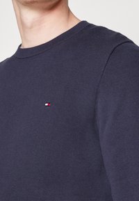 Granatowy sweter z okrągłym dekoltem wykonany z dzianiny, z małym detalem logo w kolorach czerwonym, białym i niebieskim na piersi. Gładka tekstura i klasyczny krój.