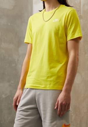 Person wearing a bright yellow Nike t-shirt and light gray shorts with an orange Nike logo, standing against a plain gray wall.