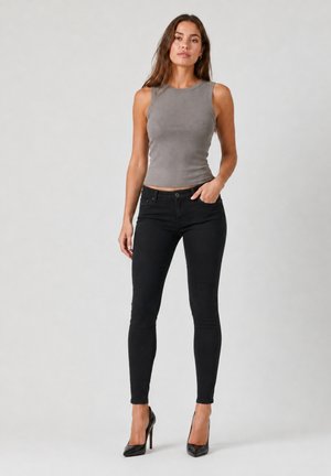 Kvinde stående iført en tætsiddende grå ærmeløs top, sorte skinny jeans og sorte højhælede sko mod en ensfarvet lys baggrund.