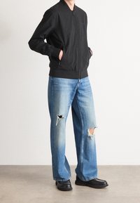 Schwarze Bomberjacke mit einem vorderen Reißverschluss, kombiniert mit hellblauen, zerfetzten Jeans mit Rissen und lockerem Schnitt. Schwarze klobige Stiefel vervollständigen den Look.