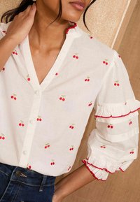 Blusa bianca con ricami di ciliegie, chiusura con bottoni, maniche a sbuffo e bordature rosse. Tessuto morbido, design casual adatto per il clima caldo.