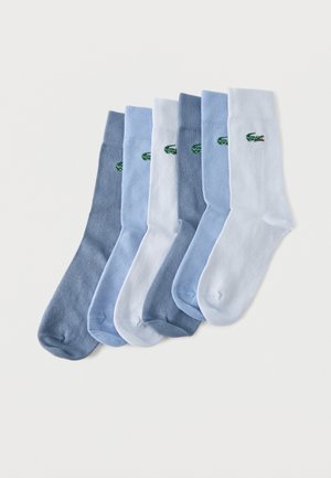 Six paires de chaussettes montantes dans des nuances de gris, bleu et blanc, chacune avec un petit logo crocodile vert près du bord, posées à plat sur une surface blanche.