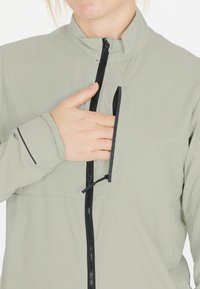 Lys grå jakke med høj krave, lynlås lukning og en brystlomme. Har sort lynlås og detaljer. Glat tekstur, let materiale.