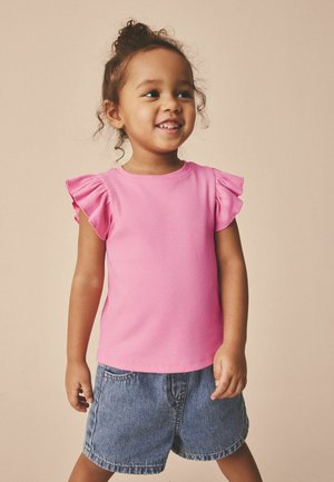 Lächendes junges Mädchen mit lockigen Haaren, das ein rosa Oberteil mit Rüschenärmeln und Jeansshorts trägt, steht vor einem beigen Hintergrund.